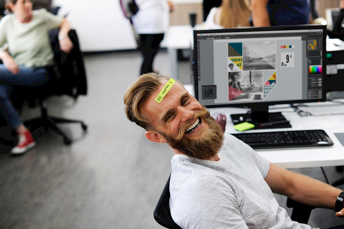 Ein junger Mann mit Bart sitzt im Büro an seinem Laptop, lacht in die Kamera und trägt ein Post-it mit der Aufschrift "be happy" auf der Stirn