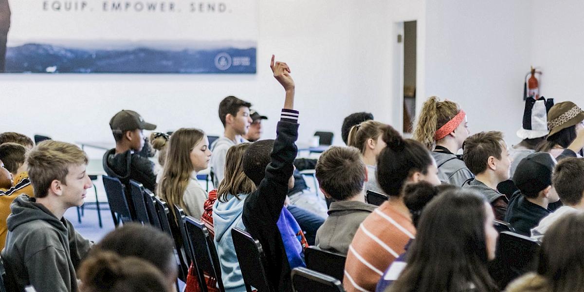 Schüler sitzen in Stuhlreihen, einer meldet sich