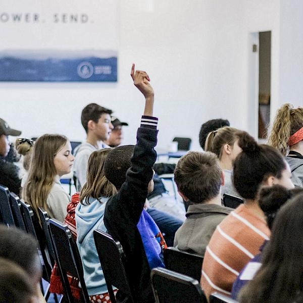 Schüler sitzen in Stuhlreihen, einer meldet sich